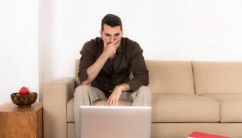 Mature man sitting on sofa looking at laptop, hand over mouth