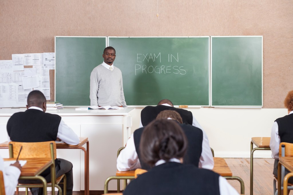 A Teacher standing by the chalk board while students write an exam. Cape Town, South Africa