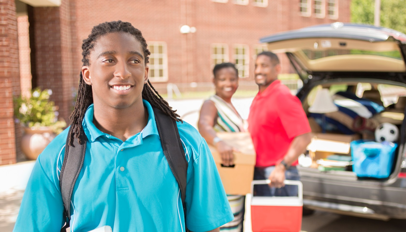 African-descent boy moves into college. Packed car, family. School campus.