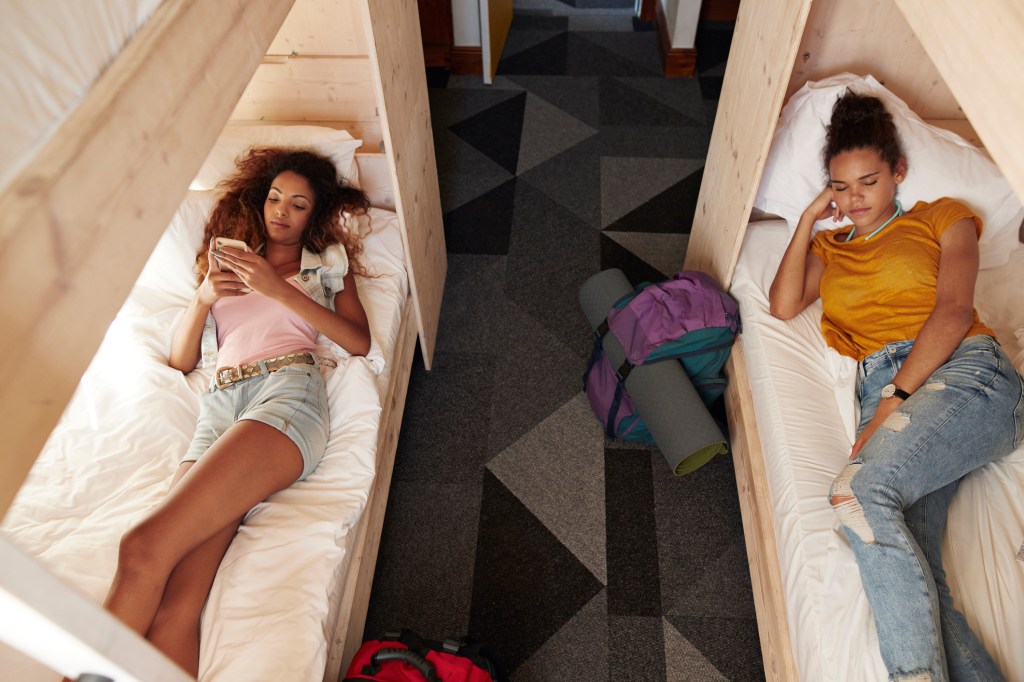 Young woman checking phone in bunk bed, roommate sleeping in the other bed