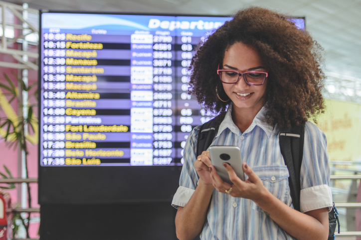 Young woman checking her boarding schedule at smartphone with airport departure time board on background