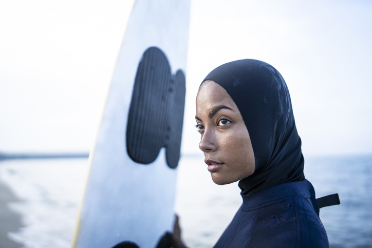 Young woman with hijab holding surfboard on beach