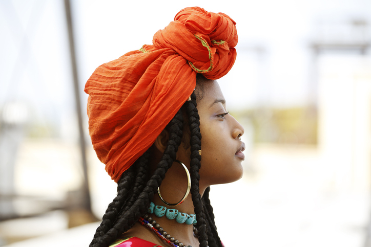 Portrait of a beautiful African American woman wearing an orange head scarf, beaded necklaces and long dreads in an outdoor setting.