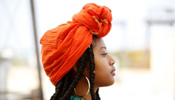 Portrait of a beautiful African American woman wearing an orange head scarf, beaded necklaces and long dreads in an outdoor setting.