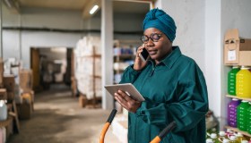 Afro female warehouse manager using a hand truck and talking on a smart phone in distribution warehouse
