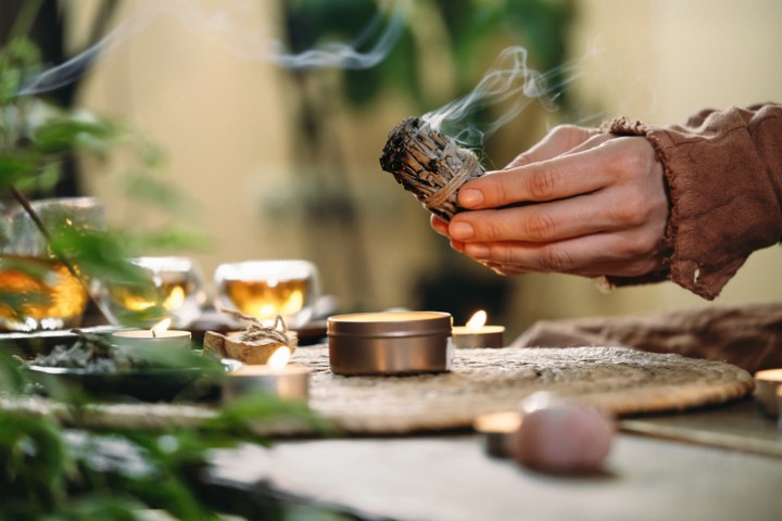 Woman Hands Burning White Sage, Before Ritual On The Table With Candles And Green Plants. Smoke Of