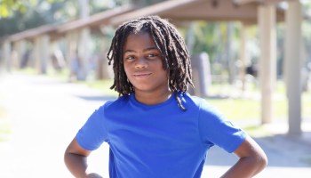 Portrait of a smiling young African American boy with locs, in an outdoor park, wearing a blue t-shirt.