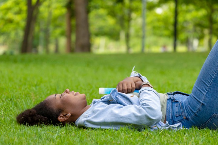 African American woman is lying down in the grass lawn inside the public park holding book in her hand during summer for reading and education concept