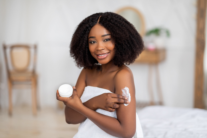 Portrait of beautiful young Afro woman enjoying morning skin care procedure, applying body cream at home