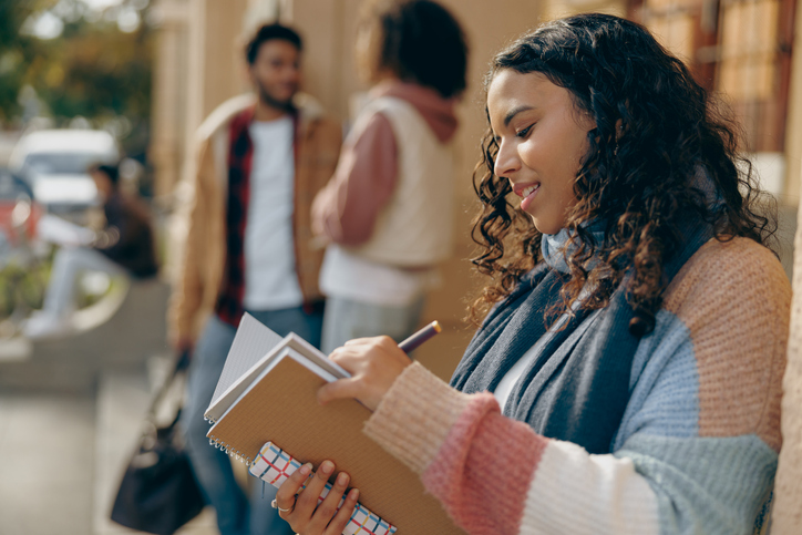 Female student making notes in notepad while standing outside of university on friends - 2023 party