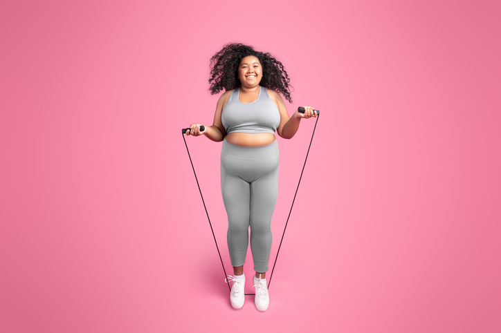 Happy african american chubby woman in sportswear jumping on jump rope, isolated on pink studio background, full length. survival mode