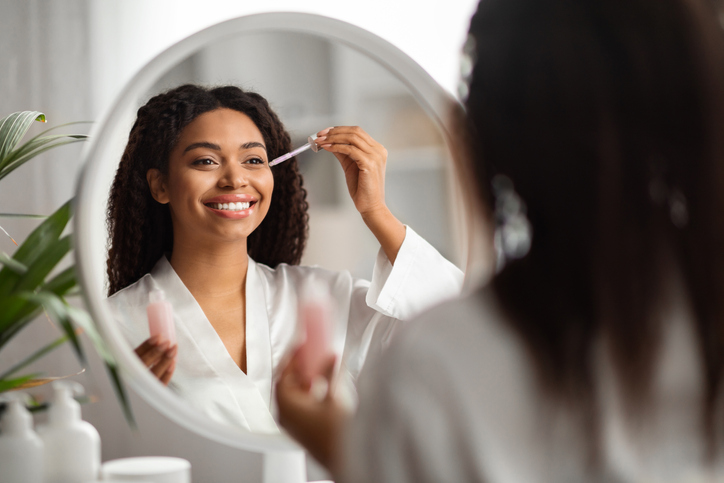 Eye Zone Care. Happy beautiful black woman applying eyes serum near mirror