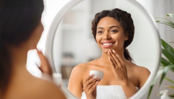 Beautiful black woman looking in mirror and applying moisturising cream on face