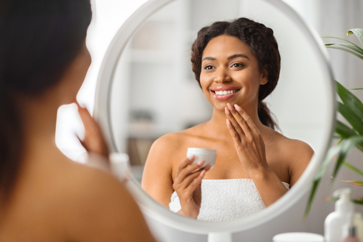 Beautiful black woman looking in mirror and applying moisturising cream on face