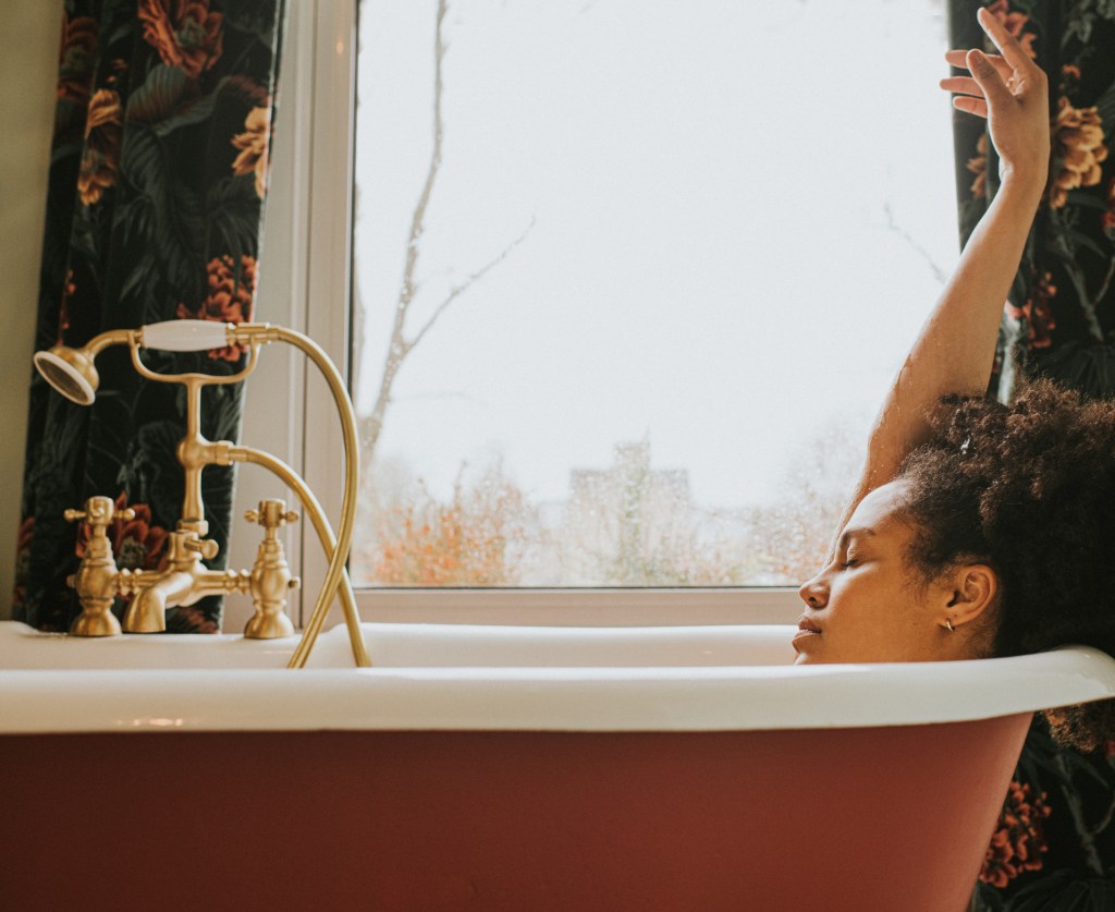 A beautiful woman bathes in a luxurious roll-top tub in front of a window, on a rainy day.