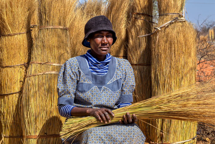African woman street vendor, crafts thatch brooms, cars on the road , in a village in Botswana , in front of thatch grass bundles for roof ,sunny day of summer