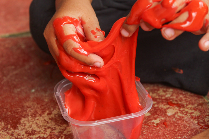 Close-up of a child's hand playing with sticky red slime. The texture of the slime looks elastic and slippery, giving it the impression of a creative sensory toy for children.