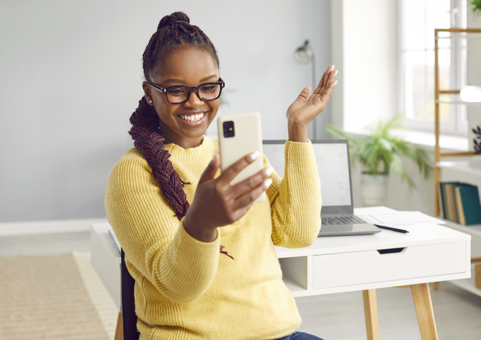 Happy smiling african-american beautiful girl looks at her mobile phone, standing in the living room against the background of desk with laptop.
