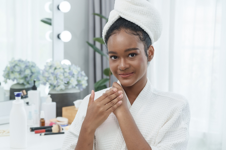 Teen african girl applying moisturizer on hands while smiling in clean beauty room wearing bathrobe and towel wrapped hair concept self care skincare wellness daily morning freshness