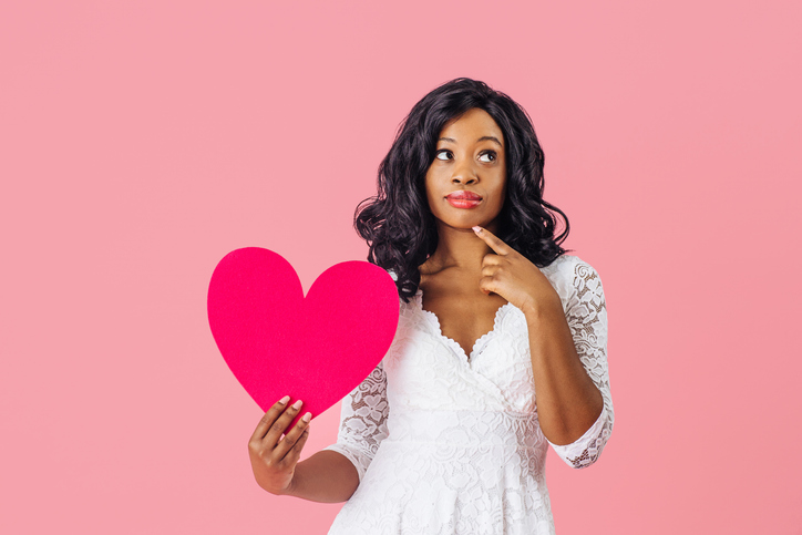 Portrait of young woman with black curly hair holding pink heart looking up and thinking, valentine' s day love and dating concept