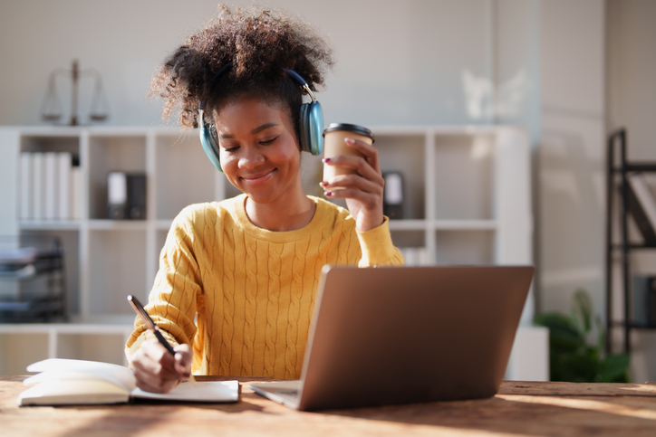 Smiling young adult woman wearing blue headphones, holding a coffee cup, and writing in a notebook while working or studying with a laptop on a wooden desk