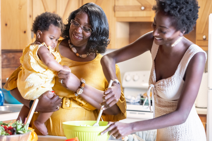 Grandmother holds baby granddaughter in kitchen while helping daughter prepare a meal for the family