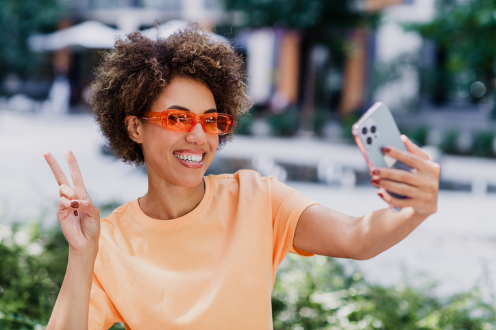 Young African American woman enjoys sunny day outdoors, taking a cheerful selfie in the city and flashing a peace sign, wearing orange shirt and red sunglasses