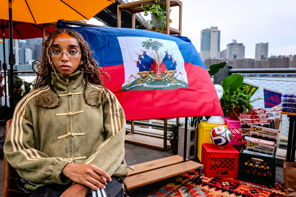 A person wearing a green jacket and glasses stands in front of a Haitian flag, surrounded by various items including a soccer ball and crates.