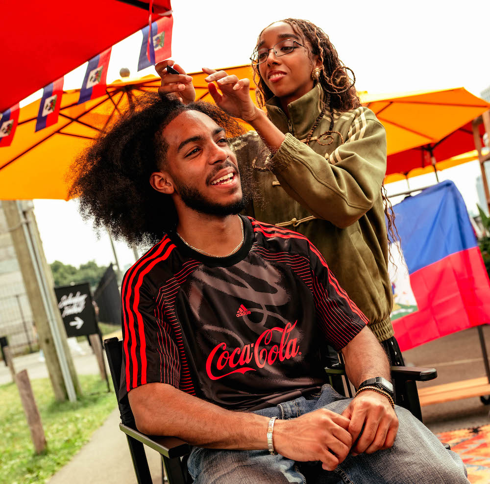 A person in a Coca-Cola jersey smiling while another person styles their hair in an outdoor setting with colorful umbrellas.
