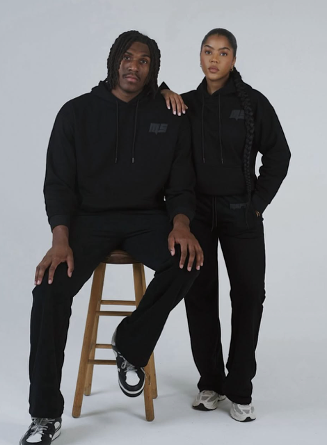 Two people wearing all-black clothing and posing together on a chair against a plain background.