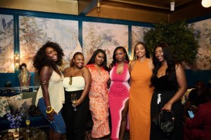 A group of six young women dressed in colorful, stylish outfits posing together in a festive setting with floral decor.