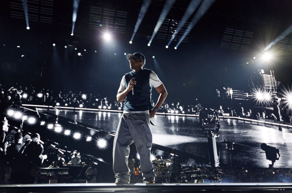 A performer on stage in a dimly lit concert venue, surrounded by a crowd of people and bright stage lights.