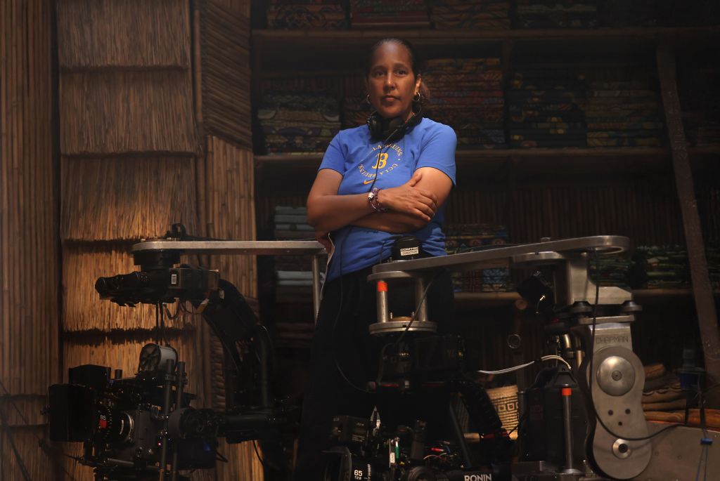 A woman in a blue t-shirt stands with her arms crossed in a room filled with film equipment, including cameras and lighting gear.