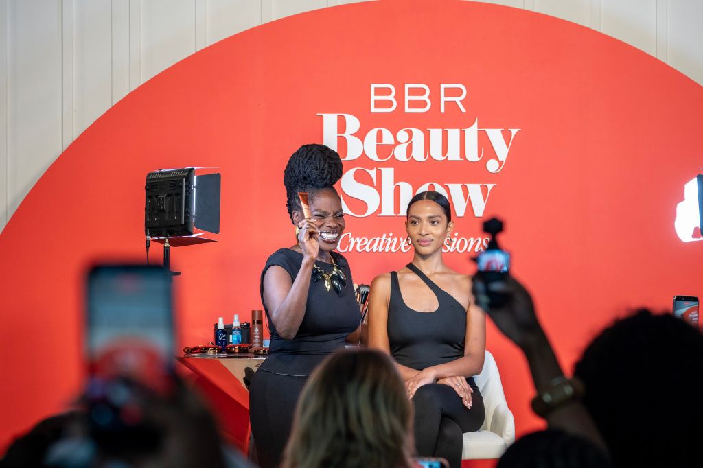 Two women on stage at the BBR Beauty Show, one speaking into a microphone and the other seated next to her.