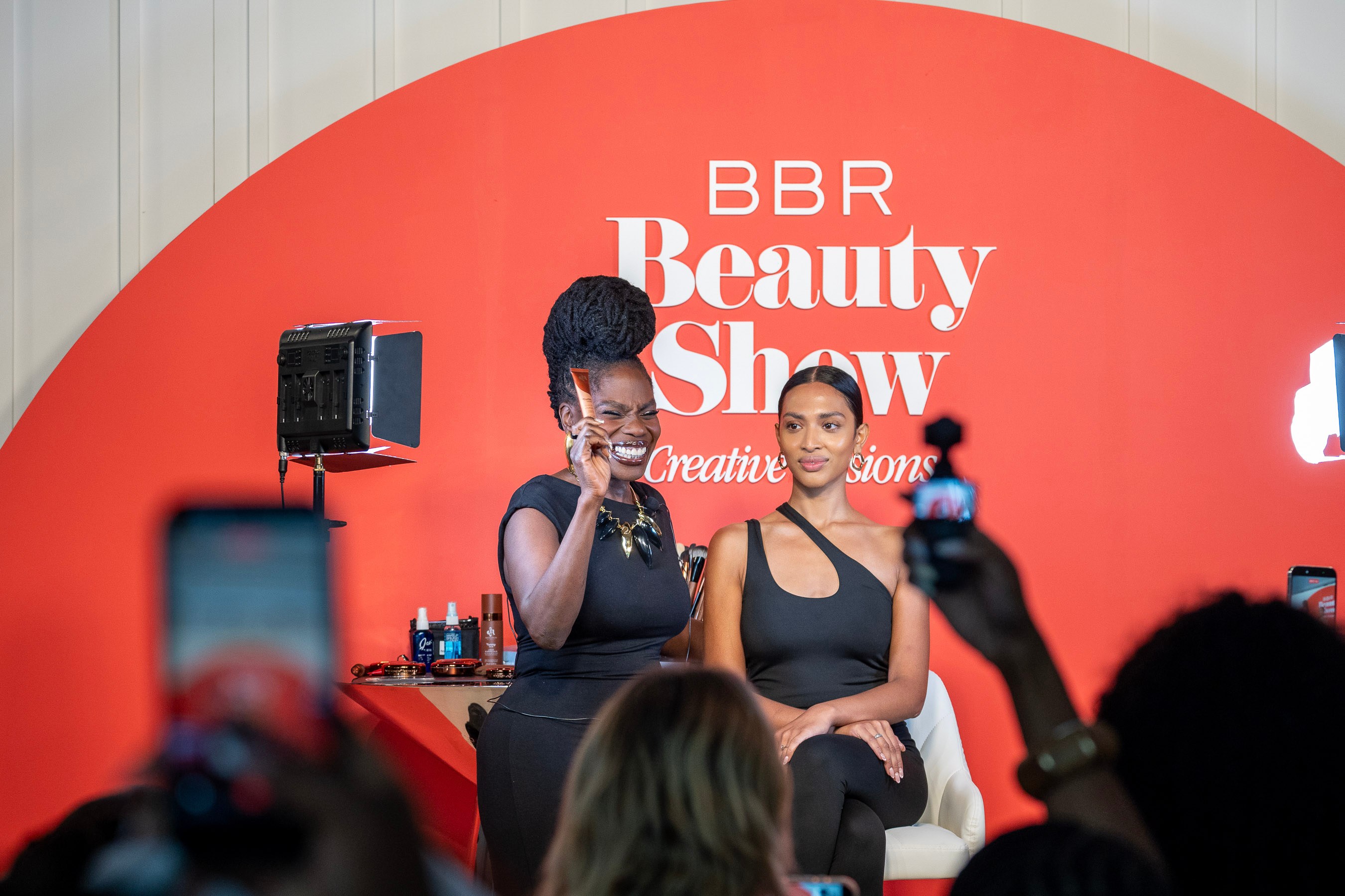Two women on stage at the BBR Beauty Show, one speaking into a microphone and the other seated next to her.