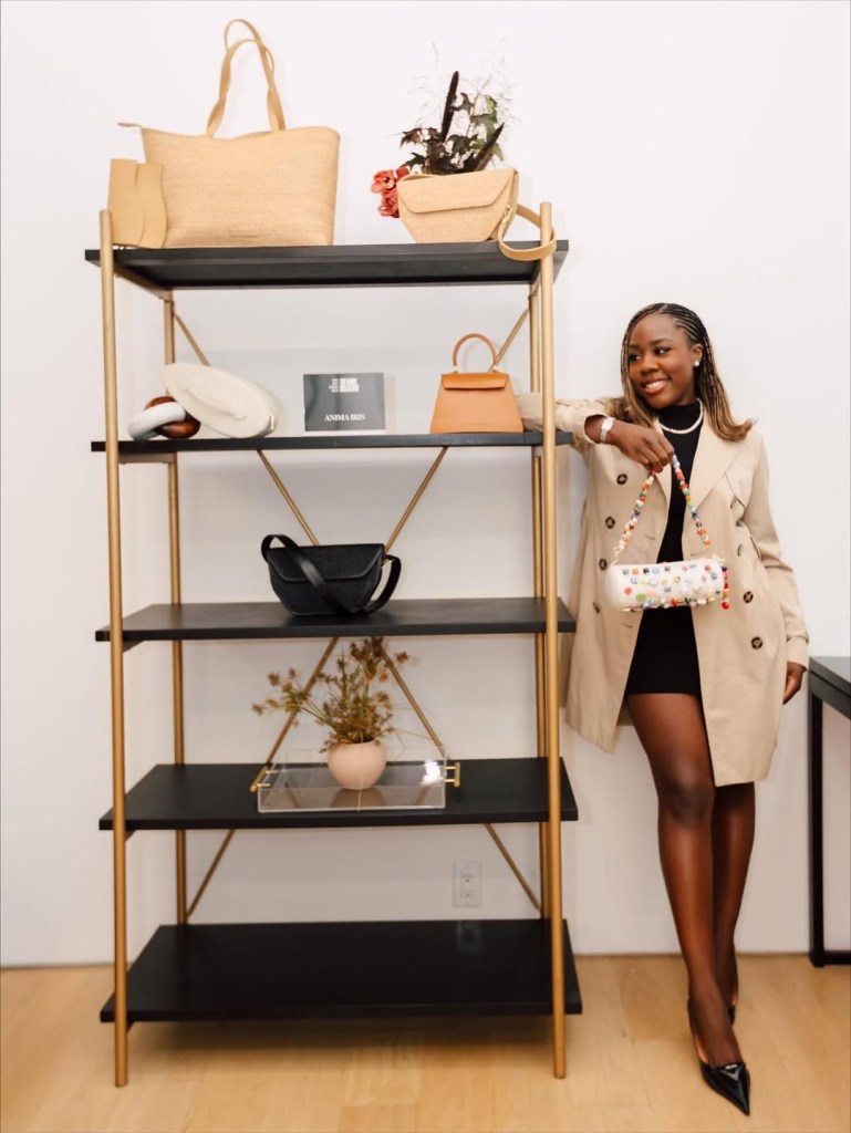 A smiling woman in a beige coat stands in front of a shelving unit displaying various handbags and accessories.