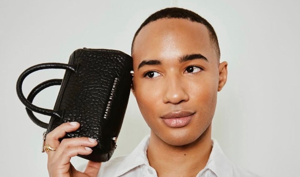 A young man wearing a white shirt and holding a black leather handbag.