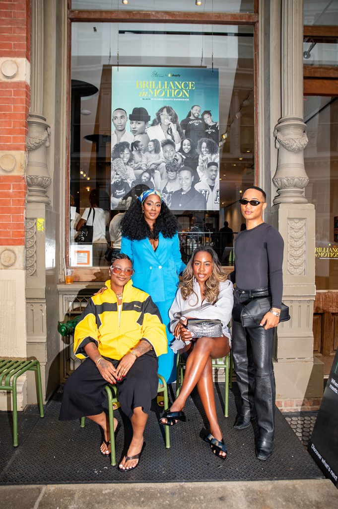A group of four people, including two women and two men, posing in front of a poster for "Brilliance in Motion", an event or exhibition. The people are dressed in colorful, stylish outfits.