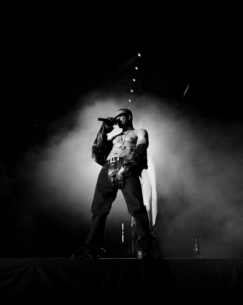 A shirtless man performing on stage, holding a microphone and surrounded by smoke.