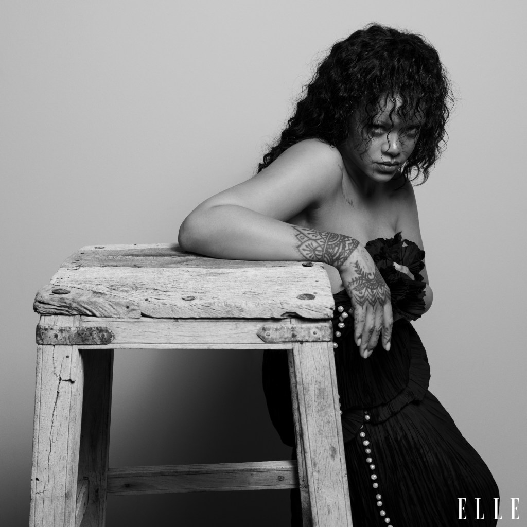 A Black and white portrait of a woman with curly hair, tattoos, and a black dress, leaning on a weathered wooden table.