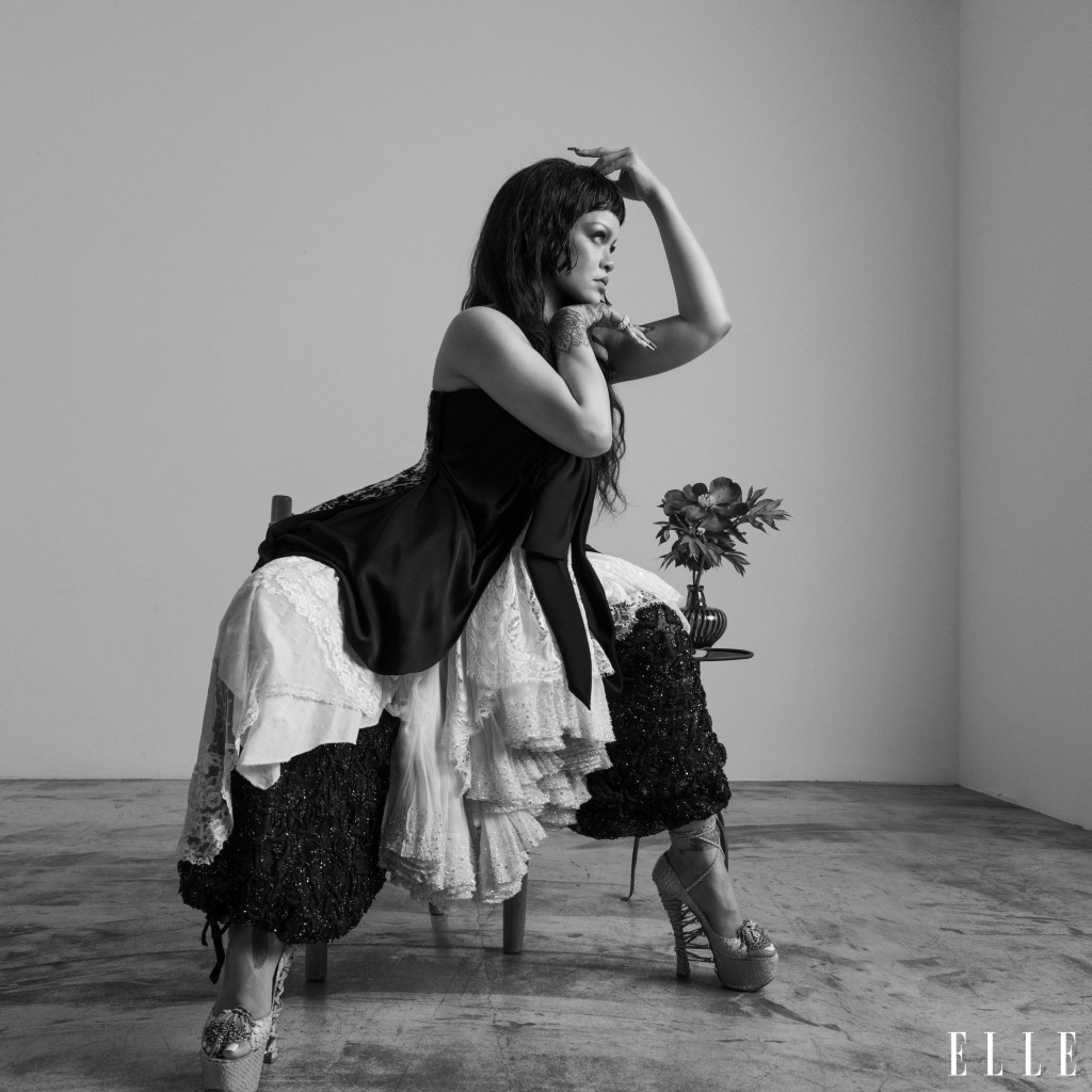 A woman in a black dress and white lace skirt stands with her arms raised, holding a bouquet of flowers in a dimly lit room.