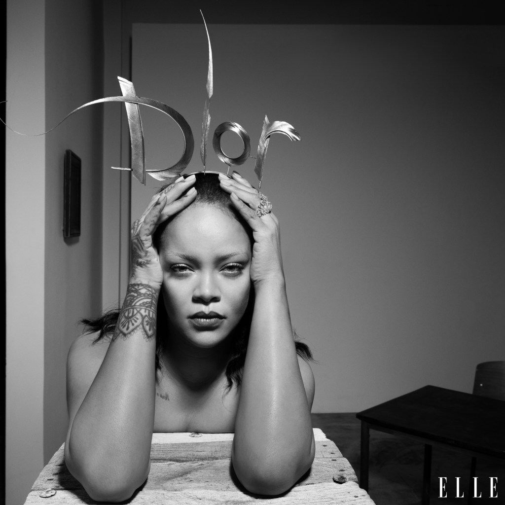 A black and white portrait of a woman with tattoos on her arms, holding scissors above her head in a contemplative pose.