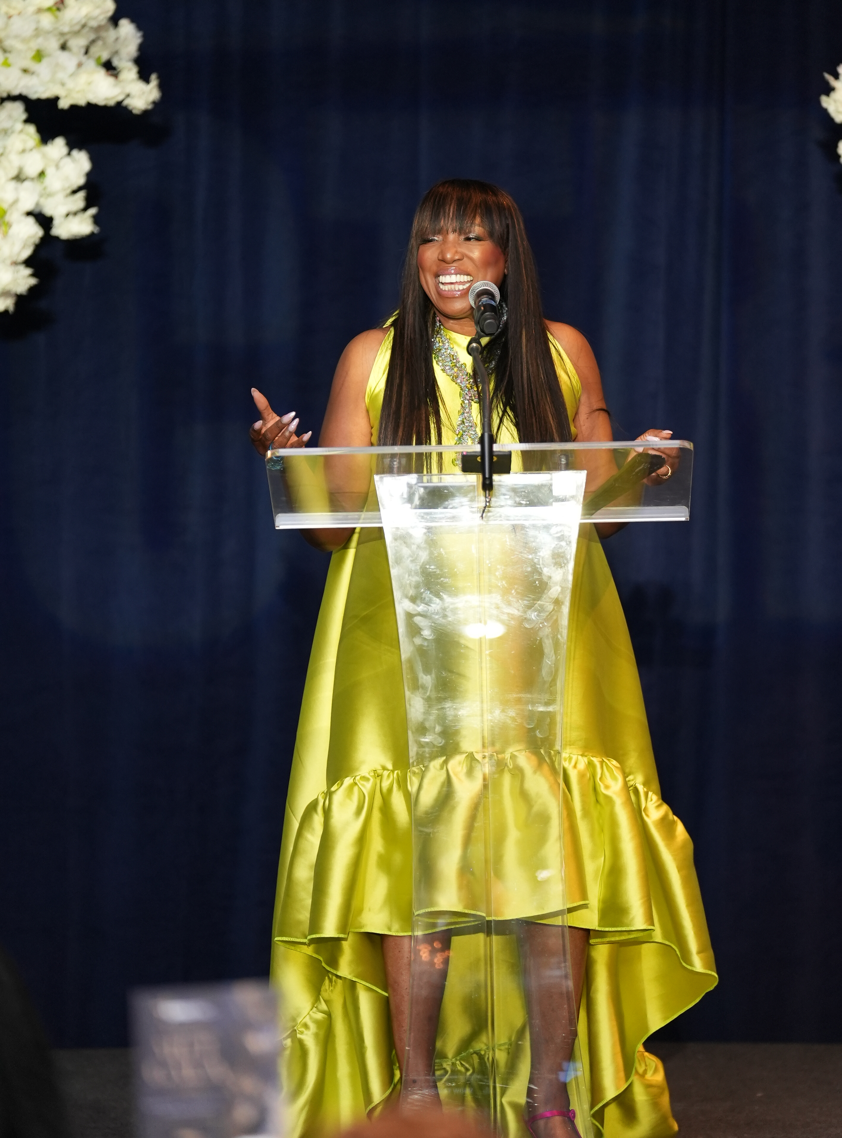 A woman in a bright yellow dress smiling and speaking into a microphone on a stage with white floral arrangements.