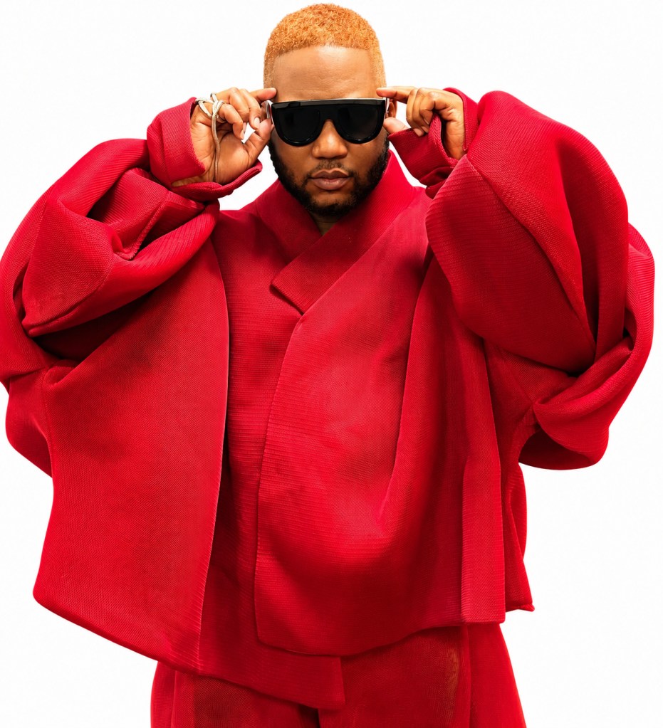 A person wearing a large, bright red coat and sunglasses, with a distinctive orange hairstyle, standing against a plain white background.