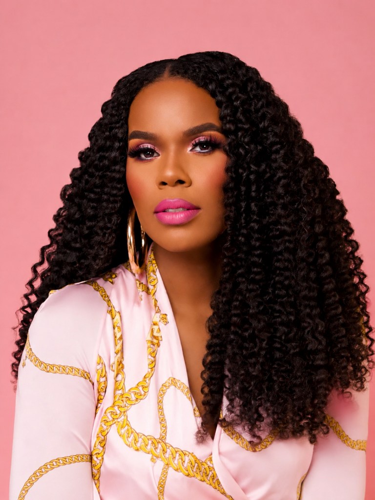 A woman with long, curly black hair wearing a white and gold patterned shirt against a pink background.