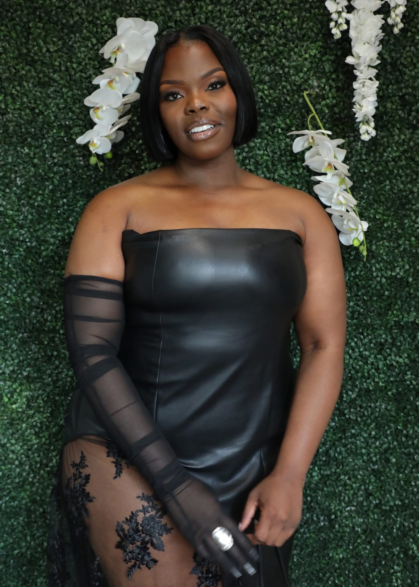 A woman wearing a black off-the-shoulder dress with sheer sleeves and a floral tattoo on her leg, standing in front of a green hedge backdrop with white orchids.
