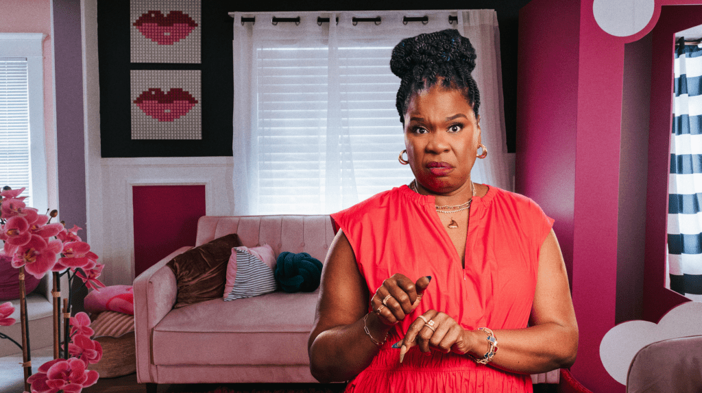 A woman in a red dress stands in a pink and black room, looking directly at the camera with a serious expression.
