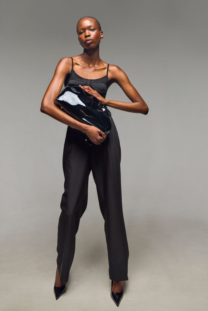 A young Black woman wearing a black tank top and pants, holding a black leather jacket, standing against a gray background.
