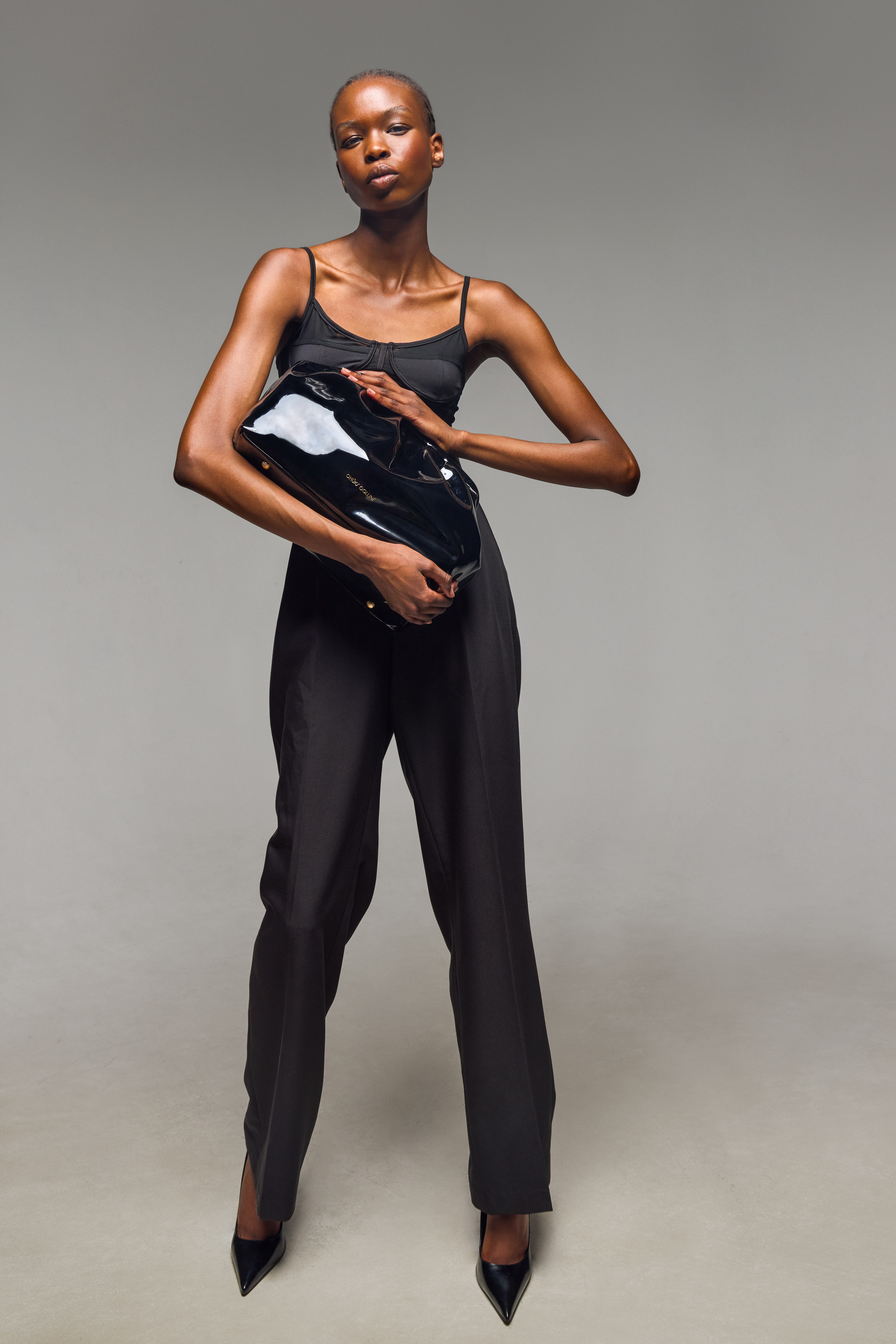 A young Black woman wearing a black tank top and pants, holding a black leather jacket, standing against a gray background.